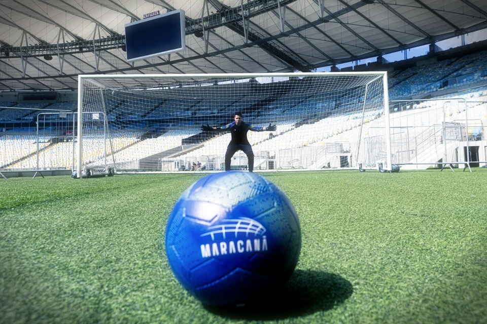 Bola oficial do Maracanã no gramado, com o goleiro posicionado no gol ao fundo.