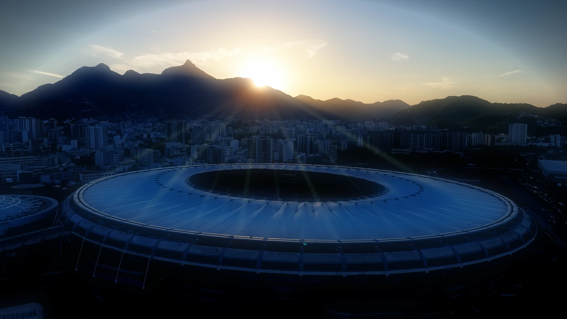 O Maracanã visto do alto no fim da tarde, com a cobertura em anel, o gramado verde e as arquibancadas vazias dentro dele, os bairros do Rio de Janeiro ao redor e a serra ao fundo.